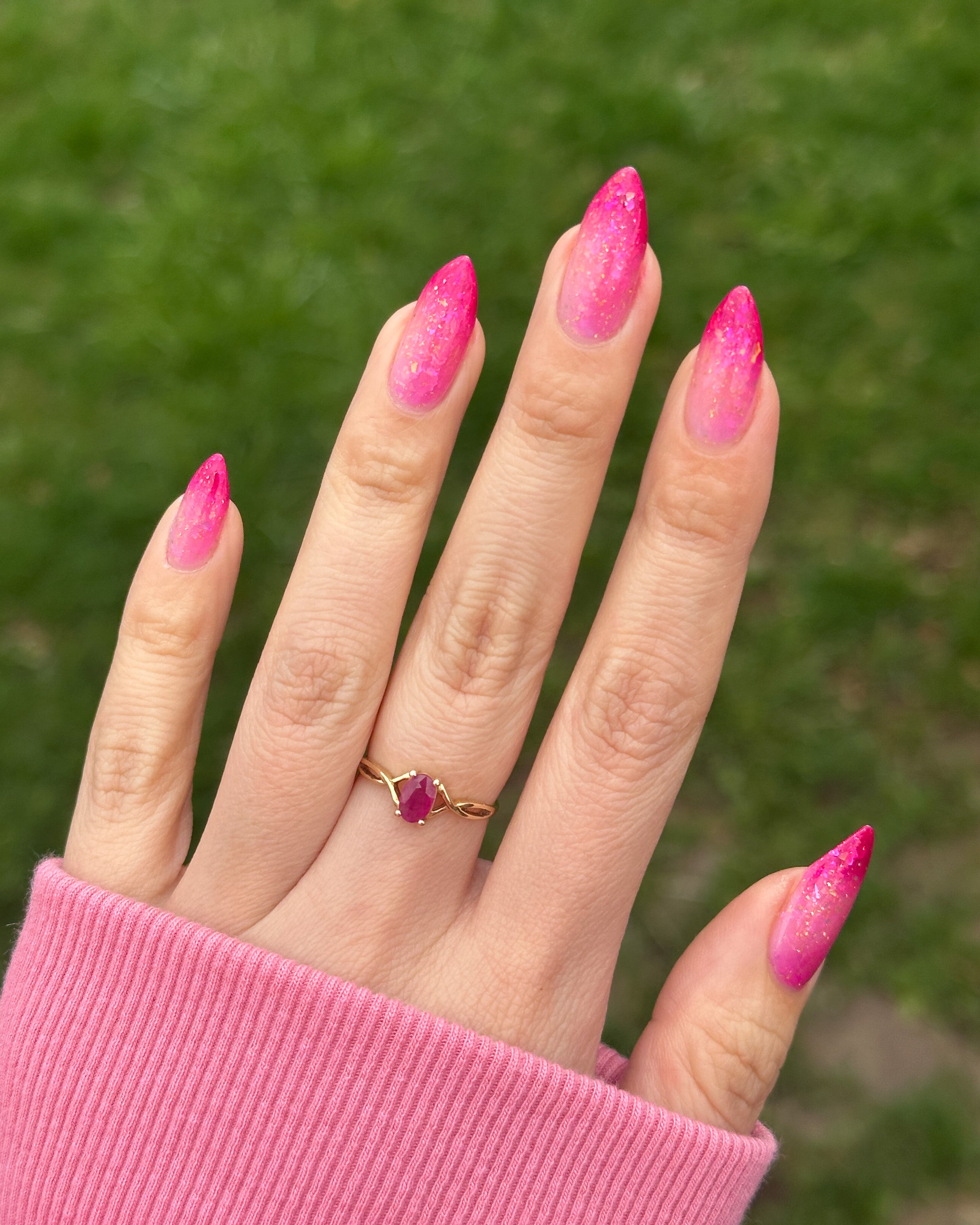 Hand with pink thermal nail polish and a gold ring with a red gemstone against a blurred green background