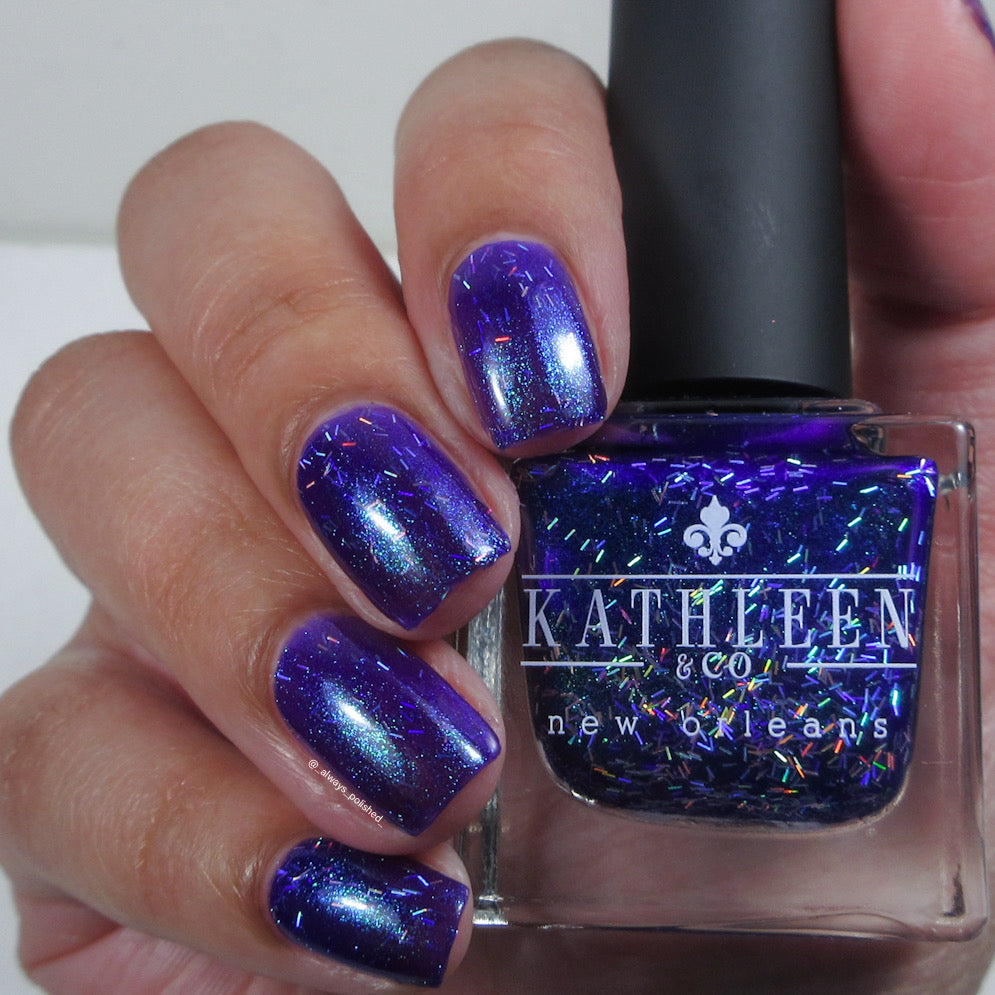 Hand with purple glittery nails holding a bottle of Kathleen &amp; Co nail polish.