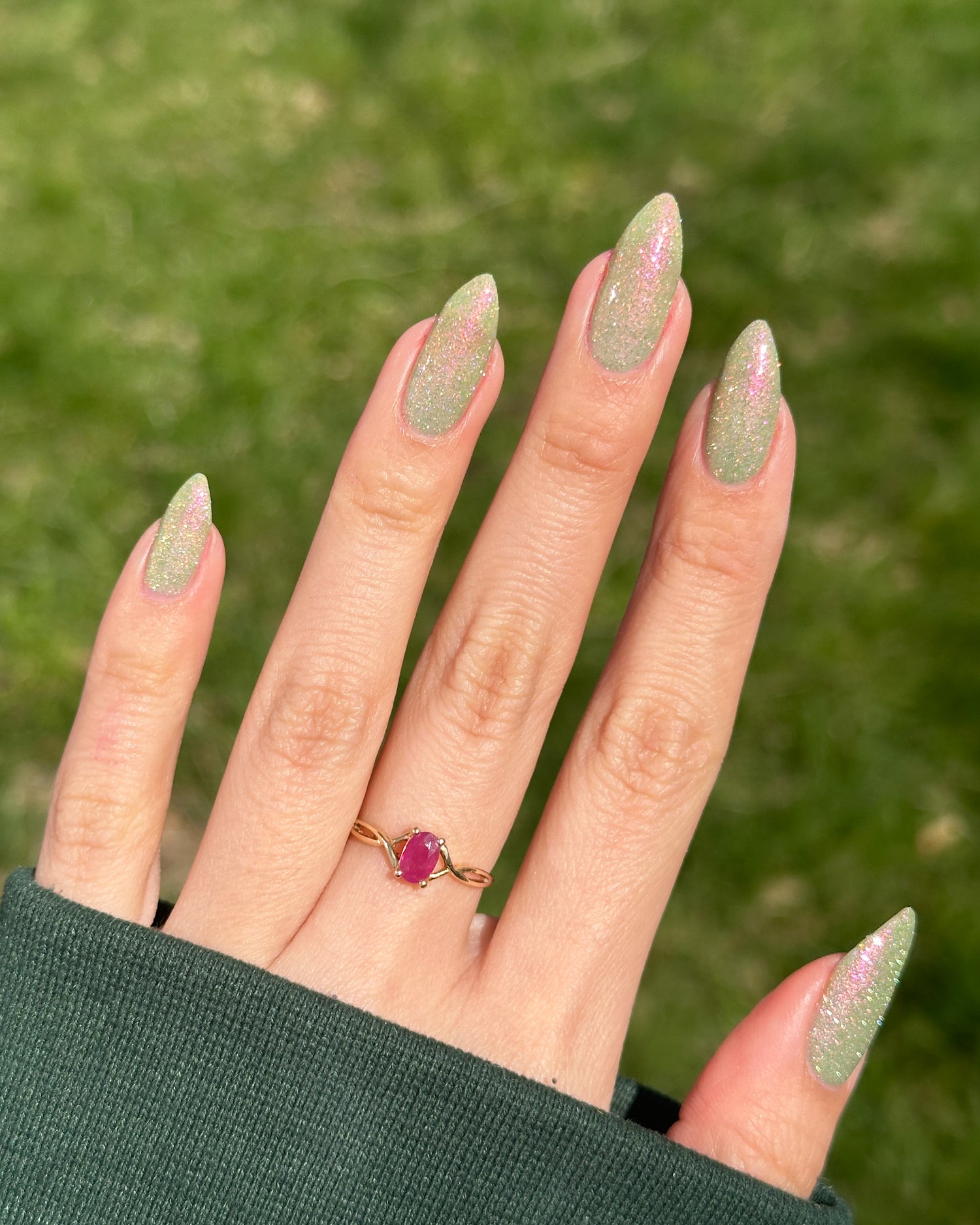 Hand with green reflective glitter nails wearing a ring on a blurred green background