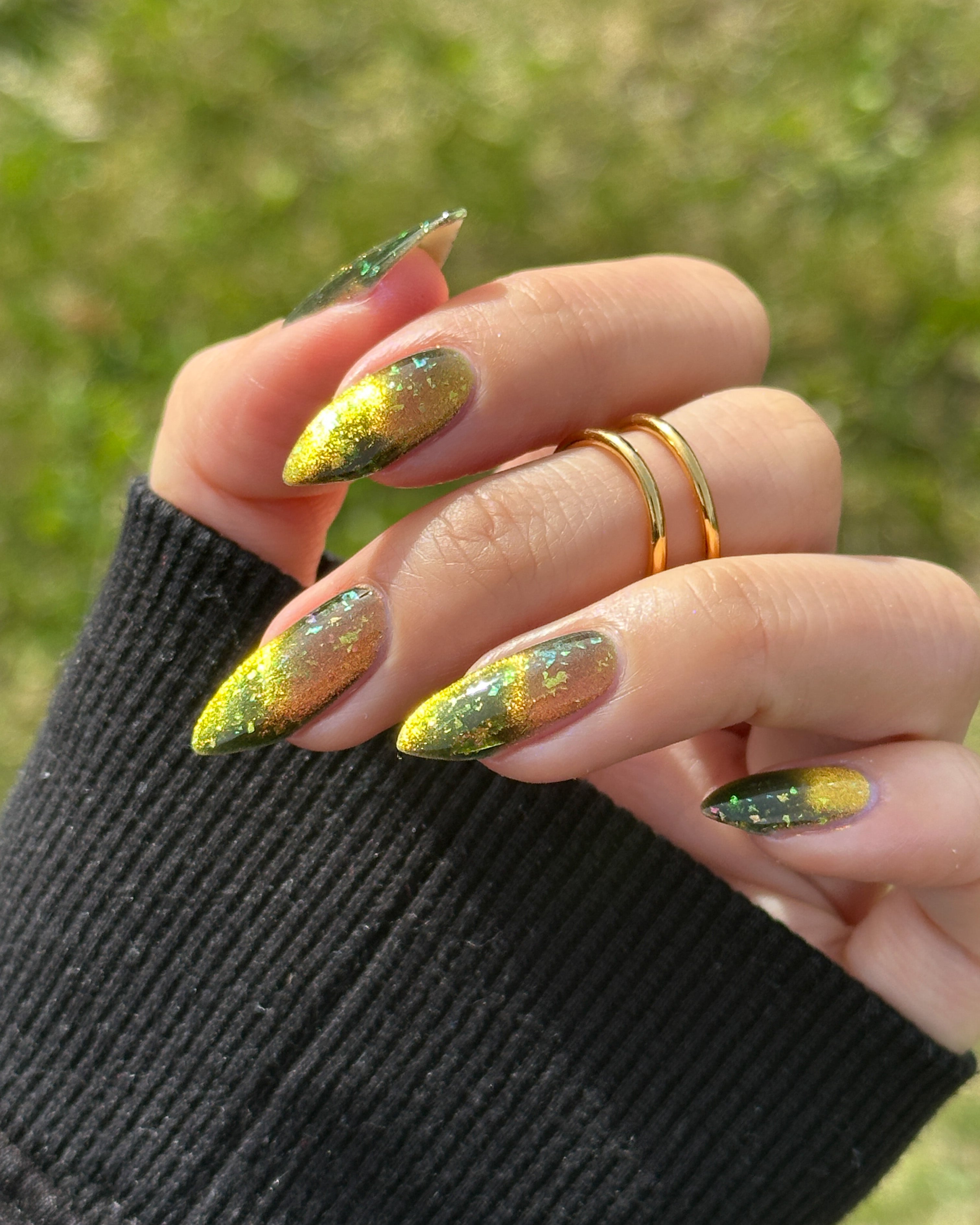 Hand with green and gold magnetic nail pollish against a blurred grass background