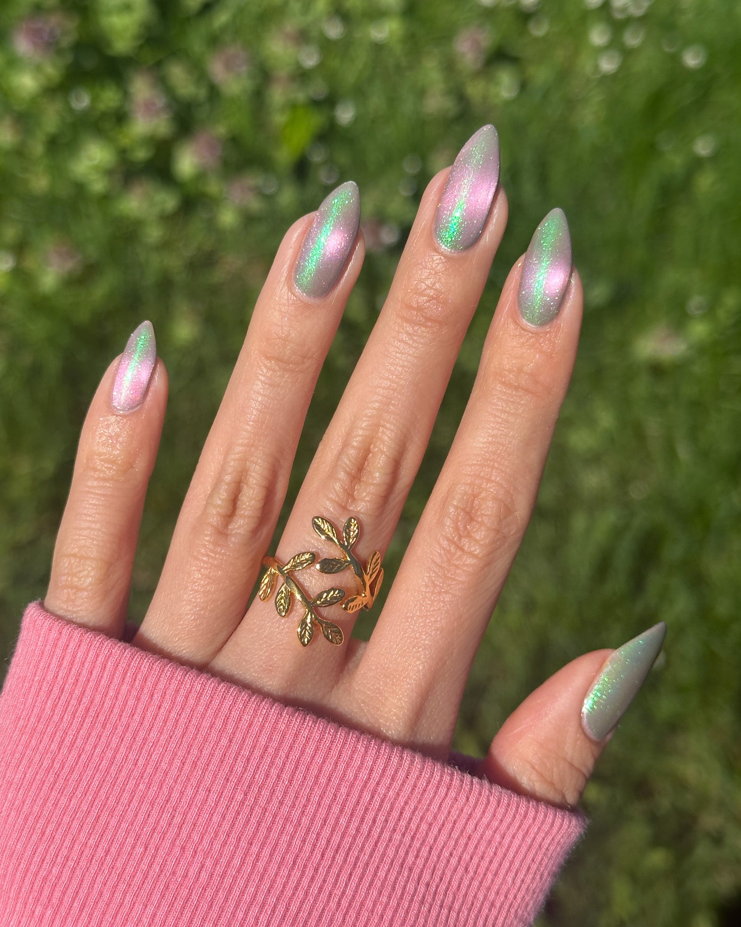 Hand with green and pink cat eye nails wearing a gold ring against a blurred green background