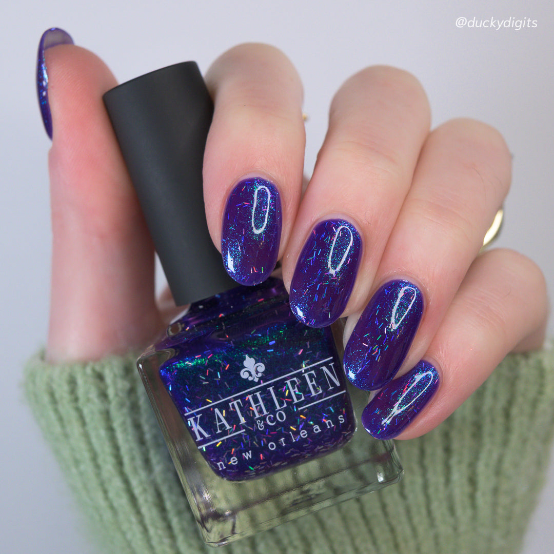 Hand holding a bottle of Kathleen &amp; Co nail polish with matching purple nails.