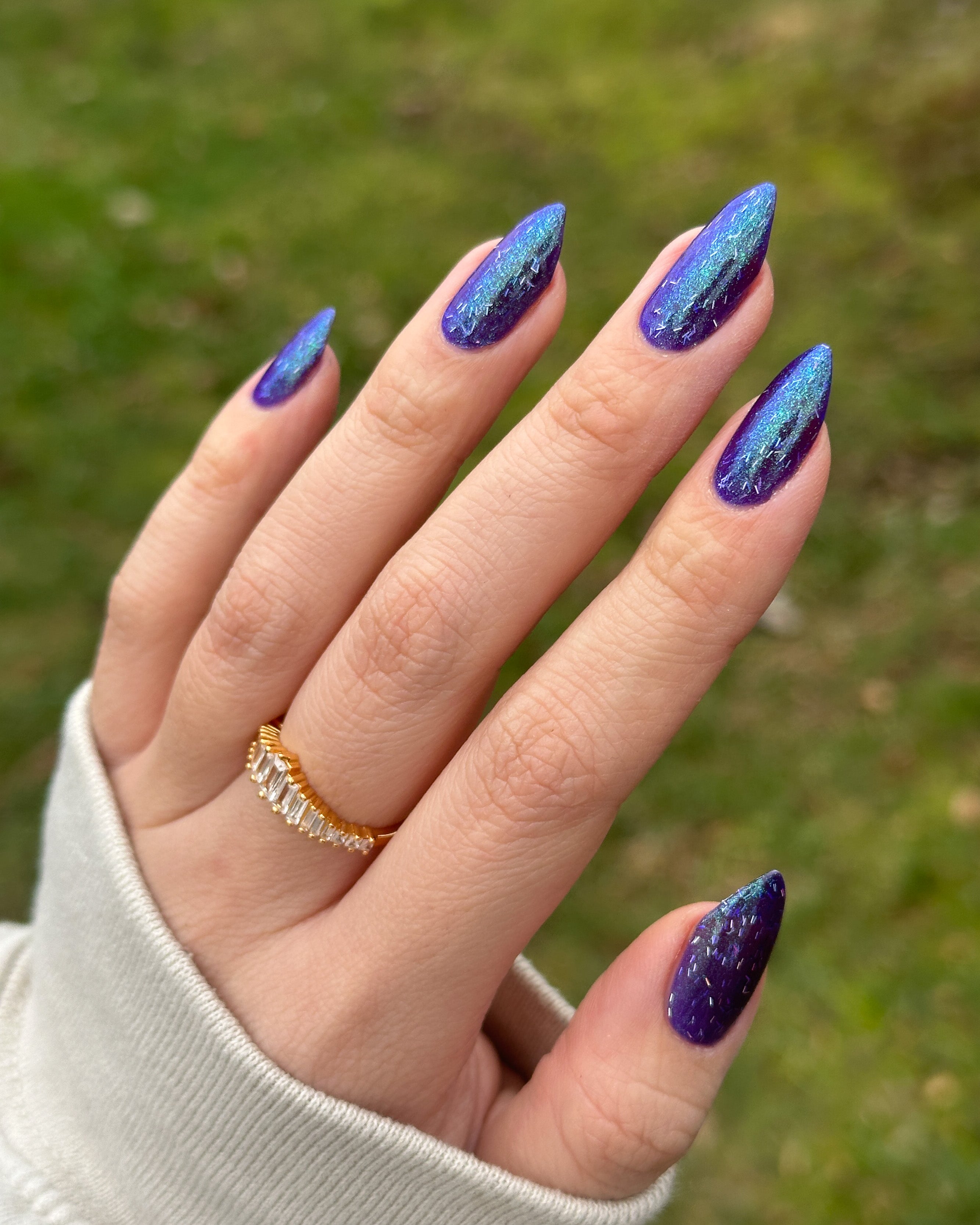 Hand with blue glittery nail polish against a blurred green background