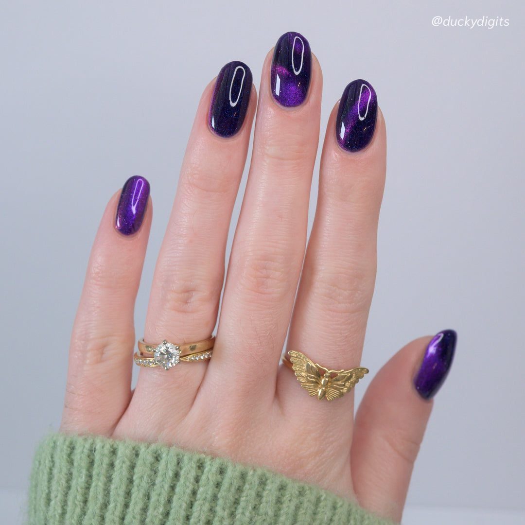 Hand with purple nail polish wearing two gold rings on a light gray background