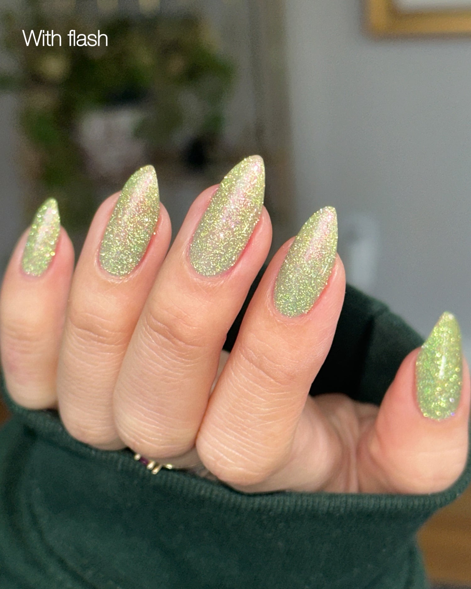 Hand with green reflective glitter nails against a blurred indoor background